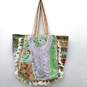 Könnte beinhalten: Eine farbenfrohe Patchwork-Tragetasche mit einer Vielzahl von Stoffmustern und -strukturen. Die Tasche zeigt eine Mischung aus floralen, geometrischen und bestickten Designs in Grün-, Lila- und Beigetönen. Weiße Quasten zieren den unteren Rand, und die Griffe sind hellbeige.