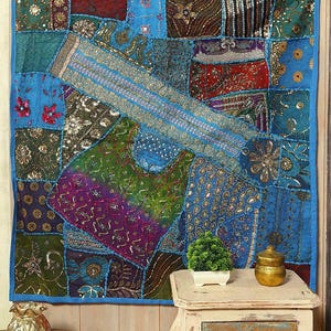 Könnte beinhalten: Ein lebendiger Patchwork-Wandbehang mit blauem Rand. Der Stoff weist eine Vielzahl von Stoffen in Blau-, Grün-, Rot- und Goldtönen auf, die mit Pailletten und aufwendigen Mustern verziert sind. Vor dem Wandbehang steht ein kleiner Tisch mit einer Pflanze und Dekorationsartikeln.
