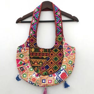 May include: A colorful, embroidered handbag with a semi-circular shape. The bag features a patchwork design with various geometric patterns in red, pink, blue, green, and yellow. The handles are a deep red with embroidered detailing, and small tassels hang from the bottom.