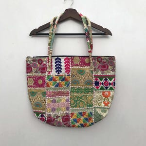 Könnte beinhalten: Eine bunte Patchwork-Tragetasche mit gestickten Blumen- und geometrischen Mustern. Die Tasche hat eine Vielzahl von Farben, darunter Rosa, Grün, Blau und Gold. Die Griffe sind aus gemustertem Stoff. Die Tasche hängt an einem Holzbügel.