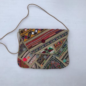 Op de afbeelding: Een kleurrijke crossbodytas met patroon en een gevlochten riem. De tas heeft een patchwork-ontwerp met verschillende geometrische en bloemmotieven in rood, geel en groen. Versieringen zijn kleine spiegels en pompons.