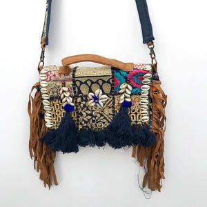Puede incluir: Bolso bandolera de estilo bohemio con bordados detallados y flecos de cuero. El bolso tiene un asa de cuero marrón y una correa azul marino. Está adornado con conchas, borlas azul marino y detalles dorados.
