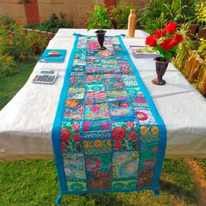 Könnte beinhalten: Ein farbenfroher Patchwork-Tischläufer mit einem lebendigen Blumendesign in Rosa, Blau und Grün. Der Läufer ist mit einem leuchtend blauen Stoff eingefasst und hat an jedem Ende Quasten. Er liegt auf einer weißen Tischdecke.