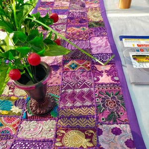 Peut inclure: Chemin de table patchwork aux tons violets, roses et dor&eacute;s. Le tissu est compos&eacute; de divers motifs brod&eacute;s et imprim&eacute;s, avec une bordure violette unie. Un vase avec des baies rouges et des feuilles vertes est visible.