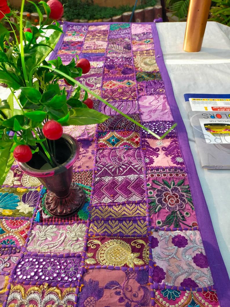 Peut inclure: Chemin de table patchwork aux tons violets, roses et dor&eacute;s. Le tissu est compos&eacute; de divers motifs brod&eacute;s et imprim&eacute;s, avec une bordure violette unie. Un vase avec des baies rouges et des feuilles vertes est visible.
