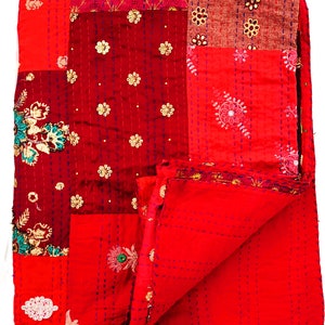 Op de afbeelding: Een rood patchwork-deken met ingewikkelde borduurwerk en een bloemenontwerp. Het deken is dubbelgevouwen en heeft een textuur.