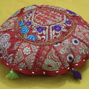 May include: Red round floor cushion with colorful embroidered floral patterns and beaded accents. The cushion has a white trim and two tassels.