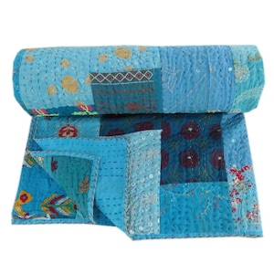 Könnte beinhalten: Eine zusammengerollte Patchwork-Quilt in Blautönen. Der Quilt zeigt verschiedene Blumen- und geometrische Muster mit sichtbaren Nähten. Die Farbpalette umfasst helle und dunkle Blautöne mit Akzenten in Gold, Rot und Grün.