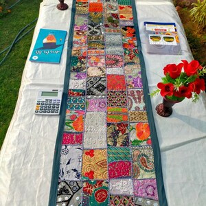 May include: A colorful patchwork table runner with a teal border. The runner is made of various patterned fabrics in shades of red, orange, green, and purple. The runner is approximately 60 inches long.