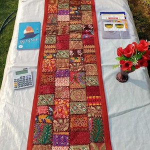 May include: A rectangular, patchwork table runner with a red border. The runner is made of various fabrics in shades of red, gold, and purple. The runner is decorated with floral and geometric patterns. A calculator and a book titled "Ideas for Life" are on the table.