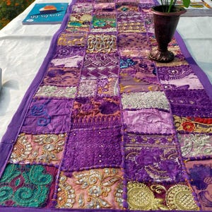 Peut inclure: Chemin de table patchwork violet, compos&eacute; de divers tissus brod&eacute;s et textur&eacute;s. Le chemin de table rectangulaire est bord&eacute; de violet uni. Des vases d&eacute;coratifs et un livre sont visibles sur la table.