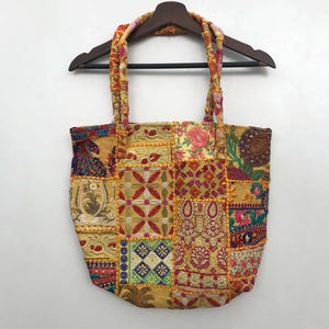 Könnte beinhalten: Eine Patchwork-Tragetasche mit einer Vielzahl farbenfroher Stoffquadrate. Die Tasche weist Blumen- und geometrische Muster in Gelb-, Rot- und Blautönen auf. Die Griffe sind geflochten und passen zum Gesamtdesign der Tasche. Die Tasche hängt an einem Holzkleiderbügel.