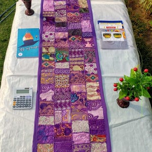 Peut inclure: Chemin de table patchwork violet, compos&eacute; de carr&eacute;s de tissus &agrave; motifs vari&eacute;s. Le chemin de table est bord&eacute; d'un tissu violet uni et comporte des glands d&eacute;coratifs aux extr&eacute;mit&eacute;s. Autres objets sur la table : une calculatrice, un livre et une petite plante.