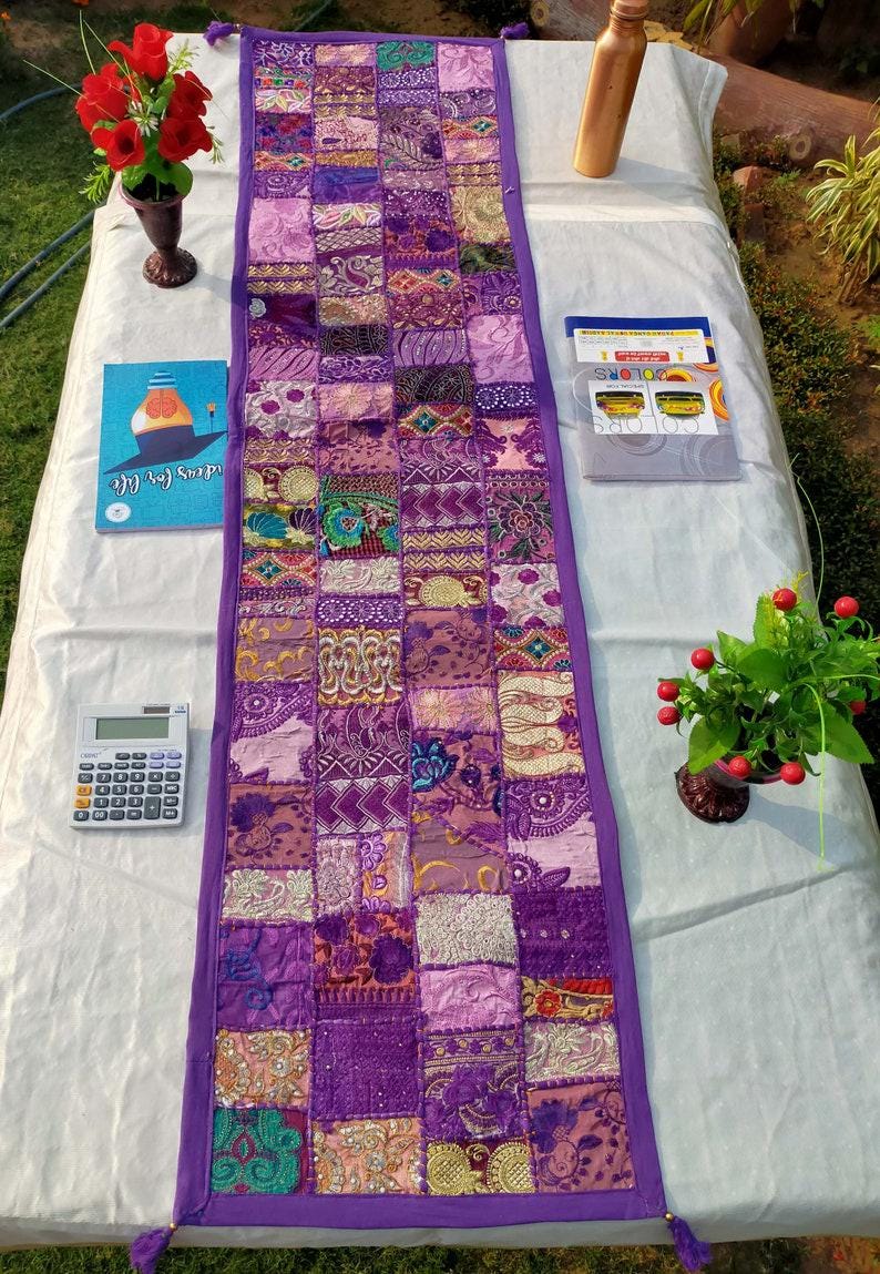 Peut inclure: Chemin de table patchwork violet, compos&eacute; de carr&eacute;s de tissus &agrave; motifs vari&eacute;s. Le chemin de table est bord&eacute; d'un tissu violet uni et comporte des glands d&eacute;coratifs aux extr&eacute;mit&eacute;s. Autres objets sur la table : une calculatrice, un livre et une petite plante.