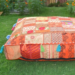 May include: A square, patchwork floor cushion in shades of orange, red, and gold. The cushion is made of various fabric pieces with floral and geometric patterns. Colorful tassels adorn the edges, adding a decorative touch. The cushion is placed on a green lawn.