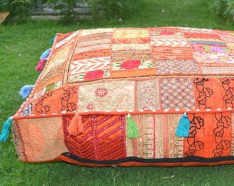 May include: A square, patchwork floor cushion in shades of orange, red, and gold. The cushion is made of various fabric pieces with floral and geometric patterns. Colorful tassels adorn the edges, adding a decorative touch. The cushion is placed on a green lawn.