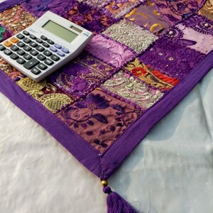 Peut inclure: Textile patchwork violet avec diverses textures et motifs. Une calculatrice blanche est pos&eacute;e sur le coin du textile. Un gland violet est suspendu au coin. Le textile est probablement un chemin de table ou une pi&egrave;ce d&eacute;corative, environ 60 cm de long.