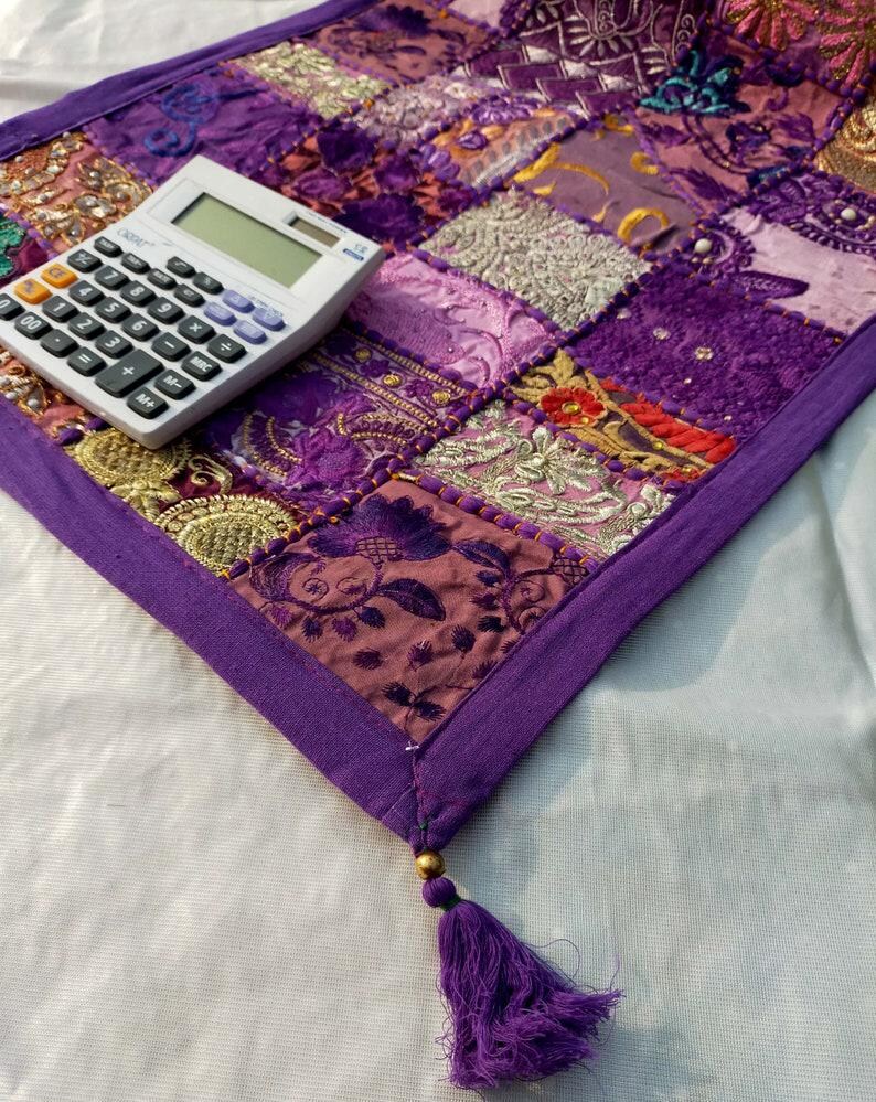 Peut inclure: Textile patchwork violet avec diverses textures et motifs. Une calculatrice blanche est pos&eacute;e sur le coin du textile. Un gland violet est suspendu au coin. Le textile est probablement un chemin de table ou une pi&egrave;ce d&eacute;corative, environ 60 cm de long.