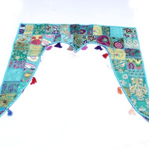 Könnte beinhalten: Ein türkisfarbener Patchwork-Volant mit hellblauem Rand und farbenfrohen Quastenakzenten. Der Volant weist eine Vielzahl von gemusterten Stoffquadraten in Blau-, Grün-, Gelb- und Rosatönen auf. Das Design ist symmetrisch.