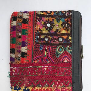 Könnte beinhalten: Eine farbenfrohe Patchwork-Geldbörse mit schwarzem Lederbesatz und Reißverschluss. Die Geldbörse zeigt eine Vielzahl von Stoffen in Rot-, Orange-, Lila- und Grüntönen mit gestickten Details und kleinen Spiegelakzenten. Das Design ist eine Mischung aus Texturen und Mustern.