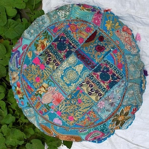 Puede incluir: Cojín de suelo redondo de patchwork en tonos turquesa, verde azulado y dorado. El cojín presenta una variedad de cuadrados de tela estampada, con diseños florales y de cachemira. Los bordes están ribeteados con un borde marrón y pequeñas borlas.