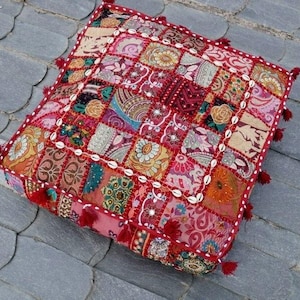 Puede incluir: Cojín de suelo cuadrado de patchwork con una vibrante mezcla de colores que incluye rojo, rosa, naranja y dorado. El cojín está adornado con pequeñas borlas rojas en cada esquina y conchas blancas a lo largo de los bordes.