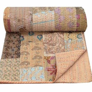 Könnte beinhalten: Eine zusammengerollte Patchwork-Steppdecke mit floralen und Paisley-Mustern in Beige-, Braun- und Rosatönen. Die Steppdecke besteht aus zusammengenähten Stoffquadraten mit sichtbaren Fäden. Sie hat einen rot-orangen Rand.