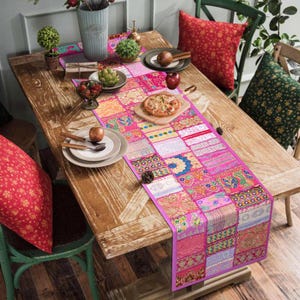 Könnte beinhalten: Ein Holztisch ist mit einem farbenfrohen Tischläufer im Patchwork-Stil in Rosa, Orange und Gold geschmückt. Der Läufer weist aufwendige Muster und einen magentafarbenen Rand auf. Teller, Obst und dekorative Kissen ergänzen die Szene.