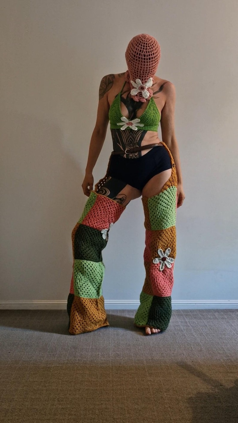 May include: A handmade crochet outfit. It includes a green bikini top with white flower accents, a matching head covering, and patchwork leg warmers in green, orange, and pink. The outfit is decorated with flower details.