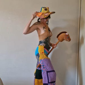 May include: A vibrant crochet ensemble including a cowboy hat, halter top, and patchwork trousers. The hat is yellow, orange, and blue. The trousers are a mix of purple, yellow, green, and floral squares. A brown stick horse is held.