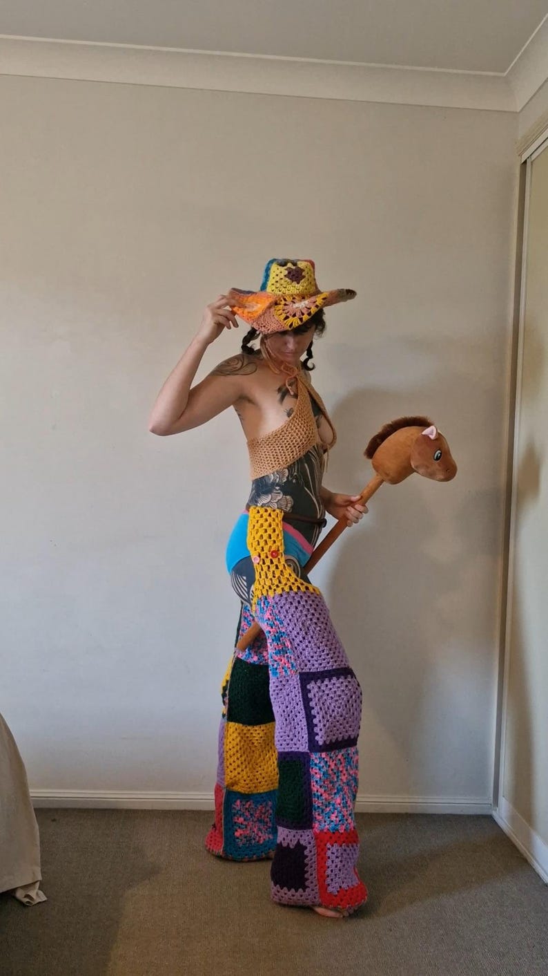 May include: A vibrant crochet ensemble including a cowboy hat, halter top, and patchwork trousers. The hat is yellow, orange, and blue. The trousers are a mix of purple, yellow, green, and floral squares. A brown stick horse is held.