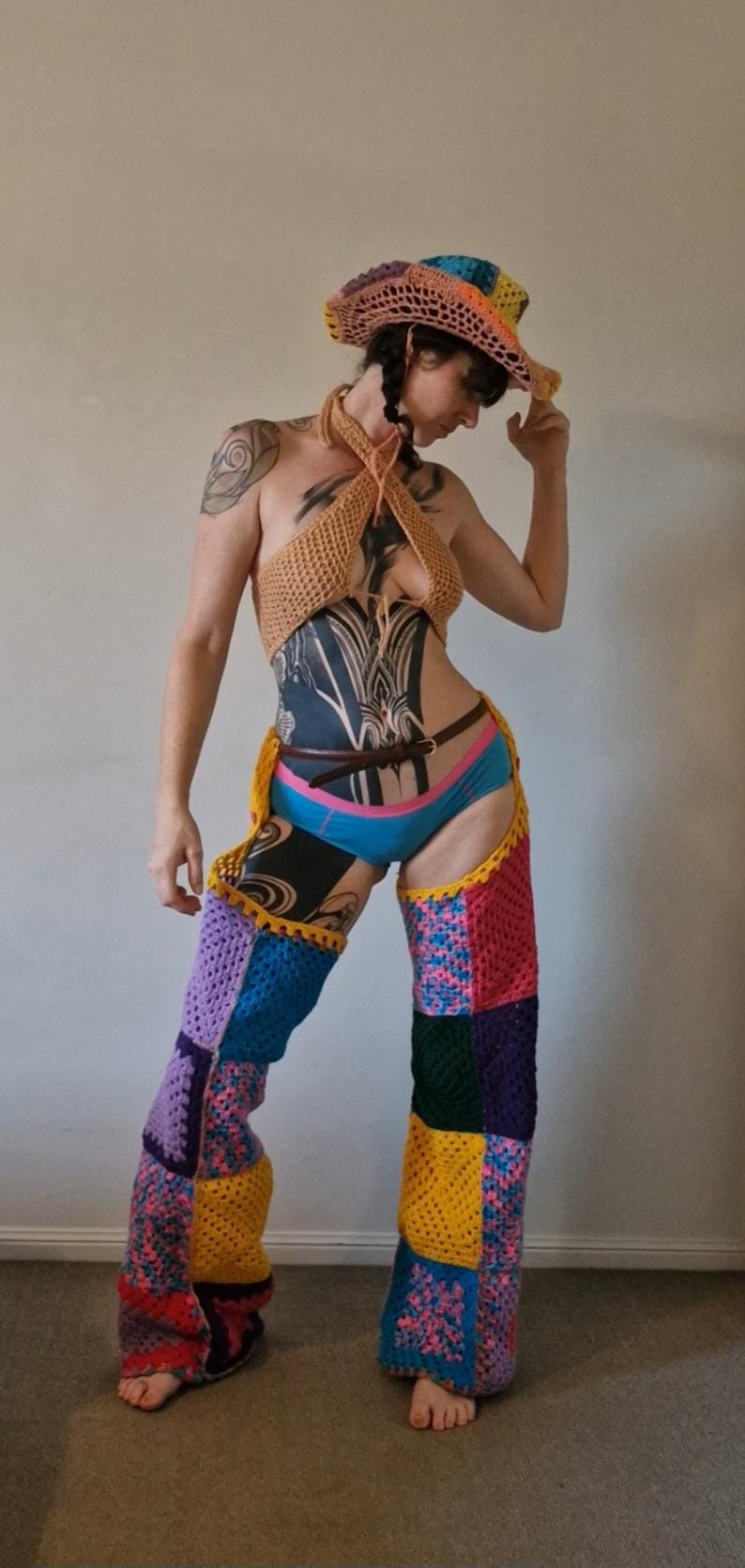 May include: A vibrant crochet ensemble comprising a halter top, chaps, and a cowboy hat. The chaps and hat are patchwork, with squares of diverse colours. The halter top is tan, and the chaps are a mix of blue, yellow, pink, and purple.