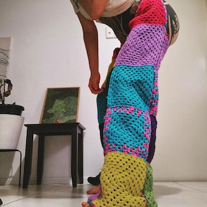 May include: A pair of colourful, patchwork crochet trousers. The trousers feature large squares of various colours, including red, purple, teal, yellow, and green, with pink accents. The open weave design is visible.