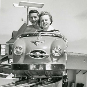 Pode incluir: Uma fotografia a preto e branco de um casal a andar num carro antigo. O carro tem um pára-choques cromado e faróis. O casal está a sorrir e parece estar a gostar do passeio.