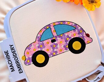 May include: Machine embroidery of a car design in a white hoop. The car is pink with purple and pink flowers, blue windows, and yellow wheels. The text "MACHINE EMBROIDERY" is on the side of the hoop. Orange flowers are in the background.