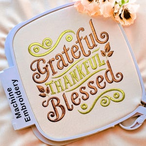 May include: A square embroidery hoop containing off-white fabric with machine-embroidered text that reads Grateful Thankful Blessed in brown and lime green thread. The design features decorative swirls and small leaf accents in a script font.