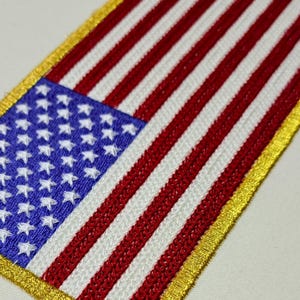 May include: Embroidered patch of the American flag, with red and white stripes and a blue canton with white stars. The patch has a gold border. The design is a detailed representation of the United States flag.