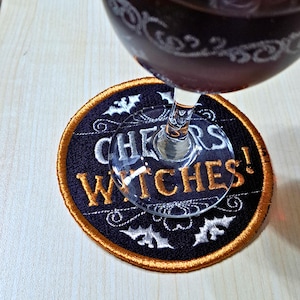May include: A circular black coaster with gold edging and the embroidered words "CHEERS WITCHES!" in white and gold. The coaster features white bat and swirl designs. A wine glass with red liquid is above the coaster.