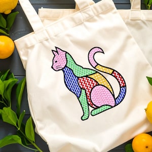 Peut inclure: Sac fourre-tout crème avec un motif de chat brodé. Le chat est divisé en sections colorées de rose, vert, bleu, jaune et rouge. Le chat est assis, la queue enroulée. Le sac a deux poignées.