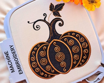 Puede incluir: Bordado a máquina de una calabaza negra con remolinos y detalles dorados, colocado en un bastidor blanco. La calabaza tiene un tallo y una hoja. El texto "MACHINE EMBROIDERY" es visible en el lateral del bastidor. Flores naranjas en el fondo.