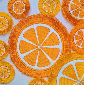 May include: Set of orange slice coasters with a fringe border. The coasters are embroidered with a white outline of an orange slice.