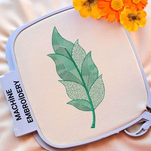 Puede incluir: Un diseño de hoja bordada en verde sobre una tela blanca. El diseño está en un aro para bordado a máquina.