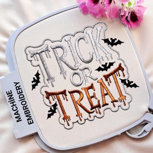 May include: Machine embroidery hoop featuring the phrase "TRICK OR TREAT" in silver and orange thread. Black bat silhouettes surround the text. The words are designed in a dripping style, ideal for Halloween.