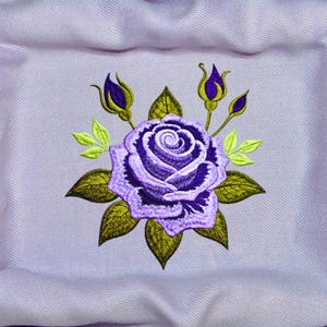 Könnte beinhalten: Ein hellviolettes Stoffquadrat mit einer zentralen Maschinenstickerei, die eine große, tiefviolette Rose mit geschichteten Blütenblättern zeigt, umgeben von dunkel olivgrünen Blättern und drei kleinen, geschlossenen violetten Rosenknospen.