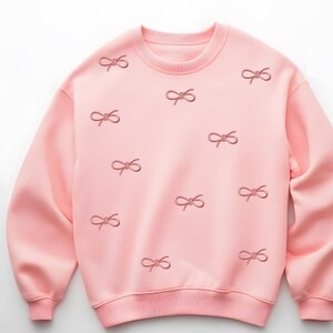 Könnte beinhalten: Ein hellrosa Sweatshirt mit Rundhalsausschnitt und gerippten Bündchen und Saum. Das Sweatshirt ist mit mehreren gestickten Schleifen in einem dunkleren Rosaton verziert, wodurch ein zartes und feminines Design entsteht. Der Stoff wirkt weich und bequem.