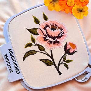 Puede incluir: Bordado a máquina de una flor rosa y oscura con hojas verdes sobre tela color crema, colocado en un bastidor blanco. El texto "MACHINE EMBROIDERY" es visible en el bastidor. Flores naranjas en el fondo.