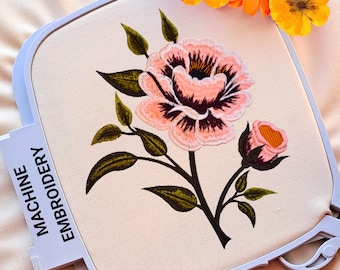Könnte beinhalten: Maschinenstickerei einer rosa und dunkelroten Rosenblüte mit grünen Blättern auf cremefarbenem Stoff, in einem weißen Stickrahmen. Der Text "MACHINE EMBROIDERY" ist auf dem Rahmen zu sehen. Orangefarbene Blumen sind im Hintergrund.