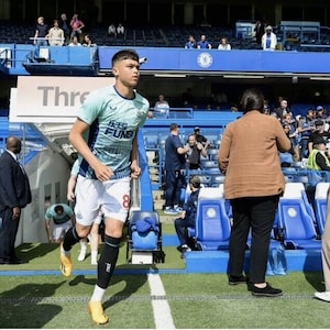 May include: A young soccer player wearing a light blue and white jersey with the text "FUN88" on the front, white shorts, and yellow socks runs onto a soccer field. The player is wearing black shin guards and yellow cleats. The player is running towards the camera. The background is a soccer stadium with blue seats and a sign that says "Three"