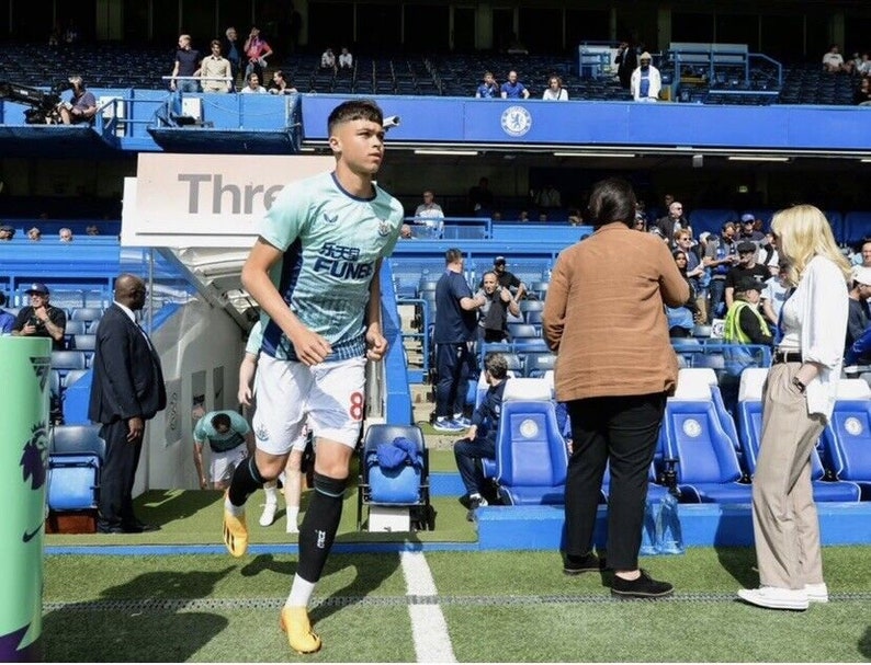 May include: A young soccer player wearing a light blue and white jersey with the text "FUN88" on the front, white shorts, and yellow socks runs onto a soccer field. The player is wearing black shin guards and yellow cleats. The player is running towards the camera. The background is a soccer stadium with blue seats and a sign that says "Three"