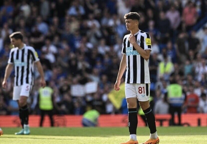May include: A young soccer player wearing a black and white striped jersey with the number 81 on the front. The player is standing on a green field with a crowd of people in the background.
